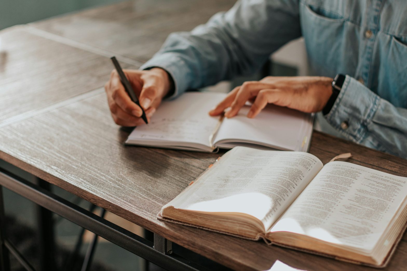 An image of a person sitting at a desk writing in a notebook and studying the Bible.